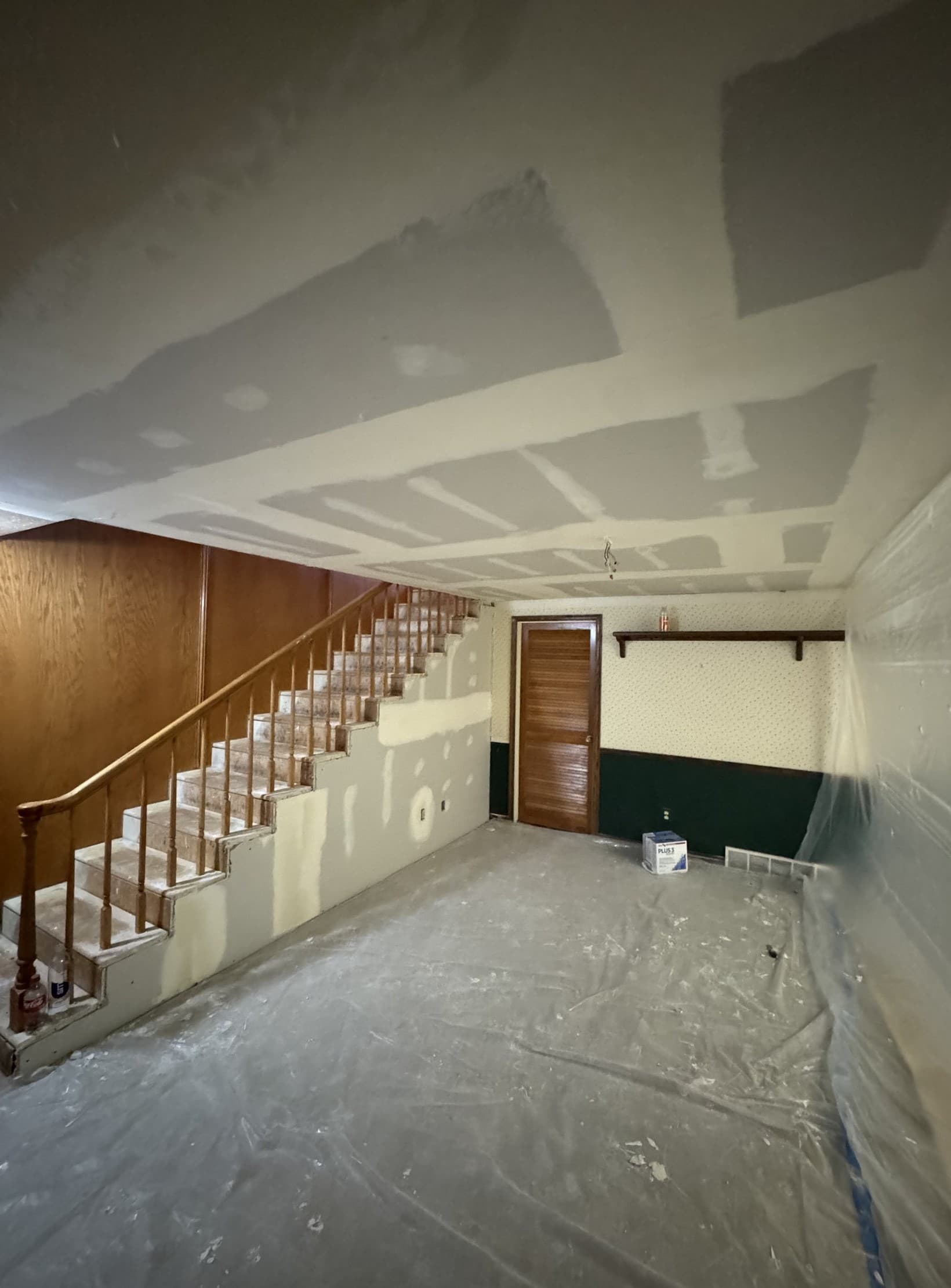 The same basement part-way through: the new ceiling board hung and every joint and screw head coated in mud waiting to be sanded, the stair wall patched and filled, and the floor and the far wall sheeted in plastic with a bucket of compound standing in the middle of the room.