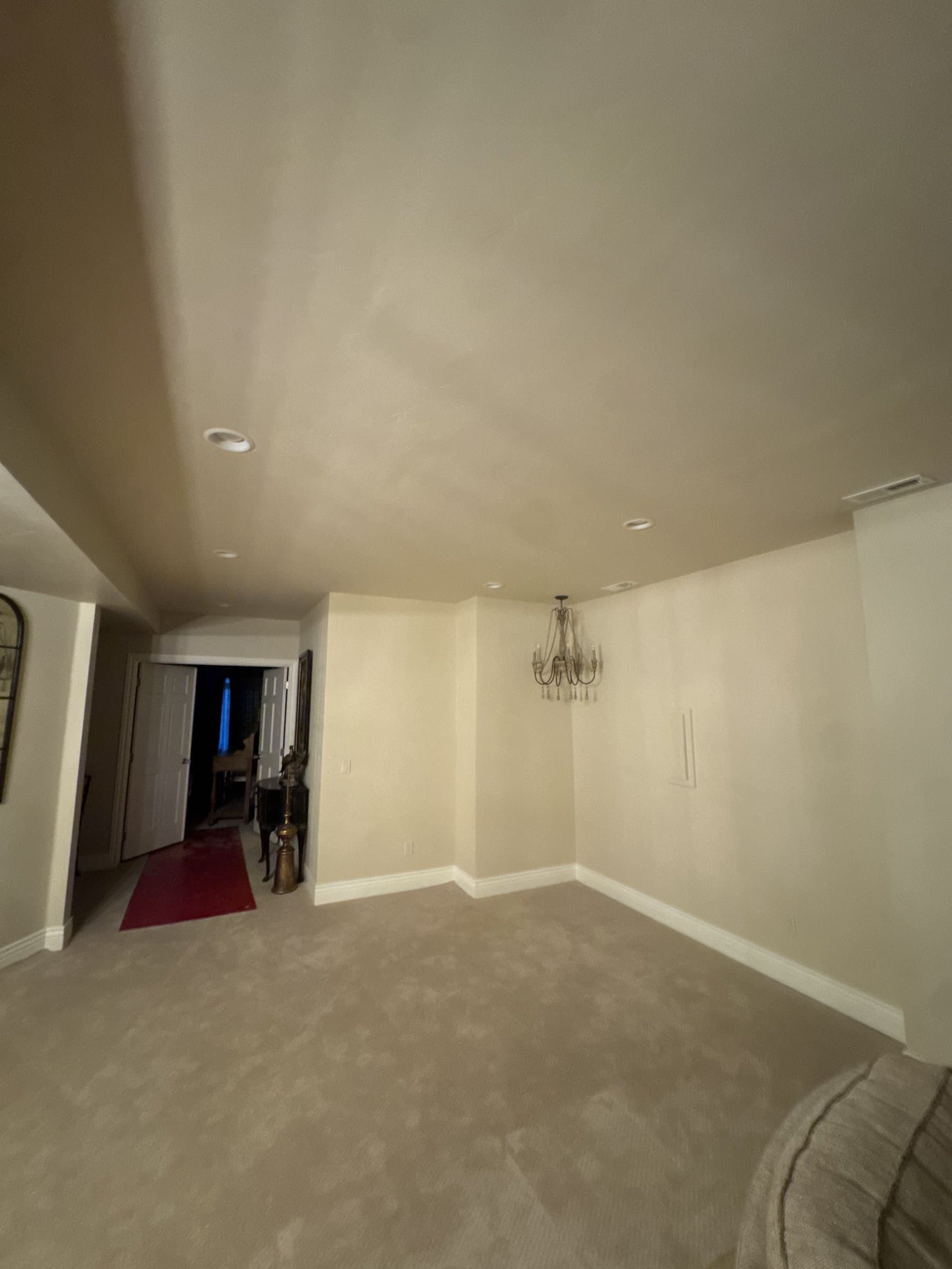 A basement sitting room with the walls and ceiling freshly rolled in a warm off-white, new white baseboard run around the corner, and a doorway through to the hall beyond.