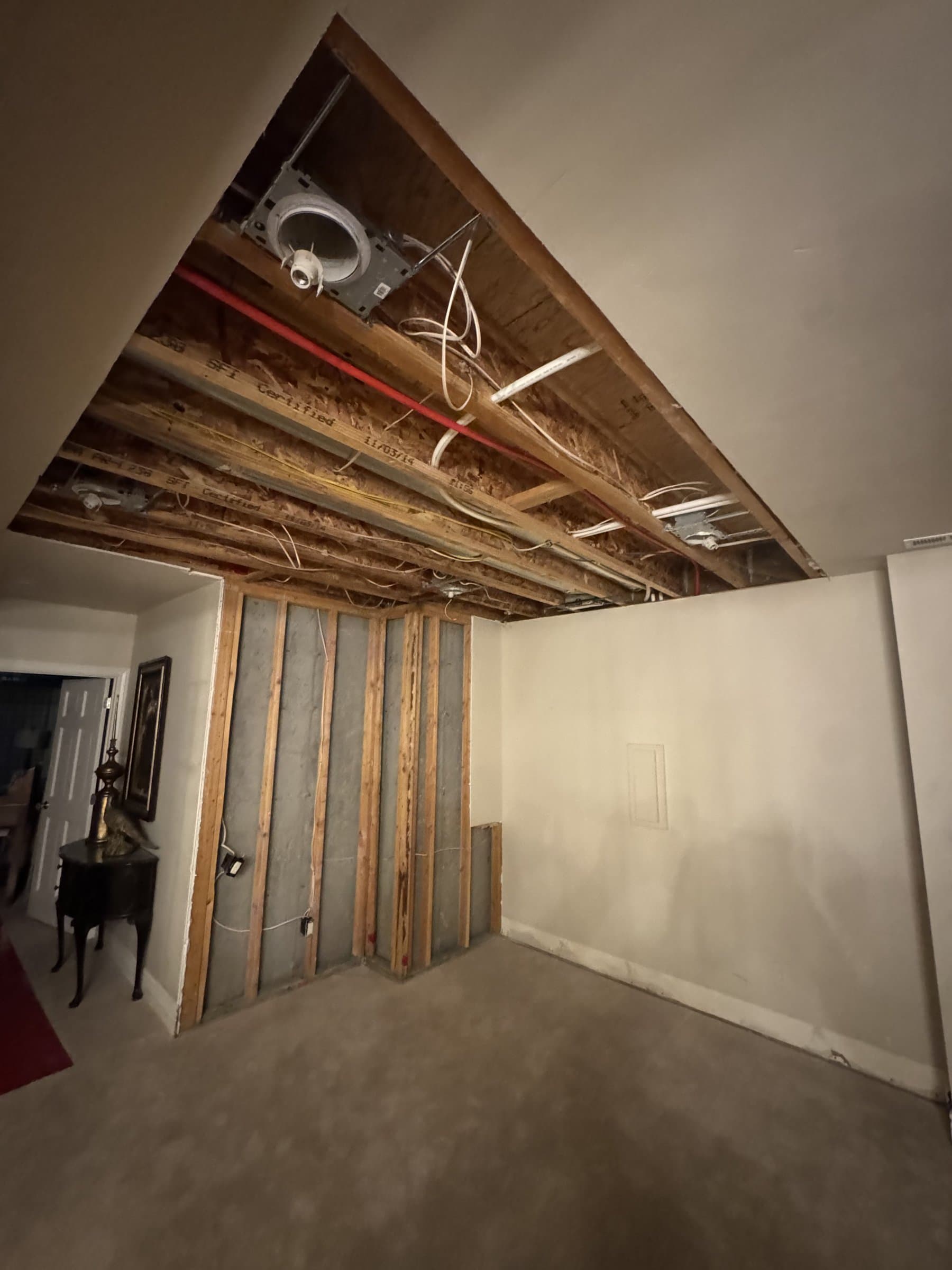 A basement with the ceiling drywall taken down to expose the joists, wiring and pipework above, and one wall stripped back to studs against the concrete, with the old carpet still on the floor.