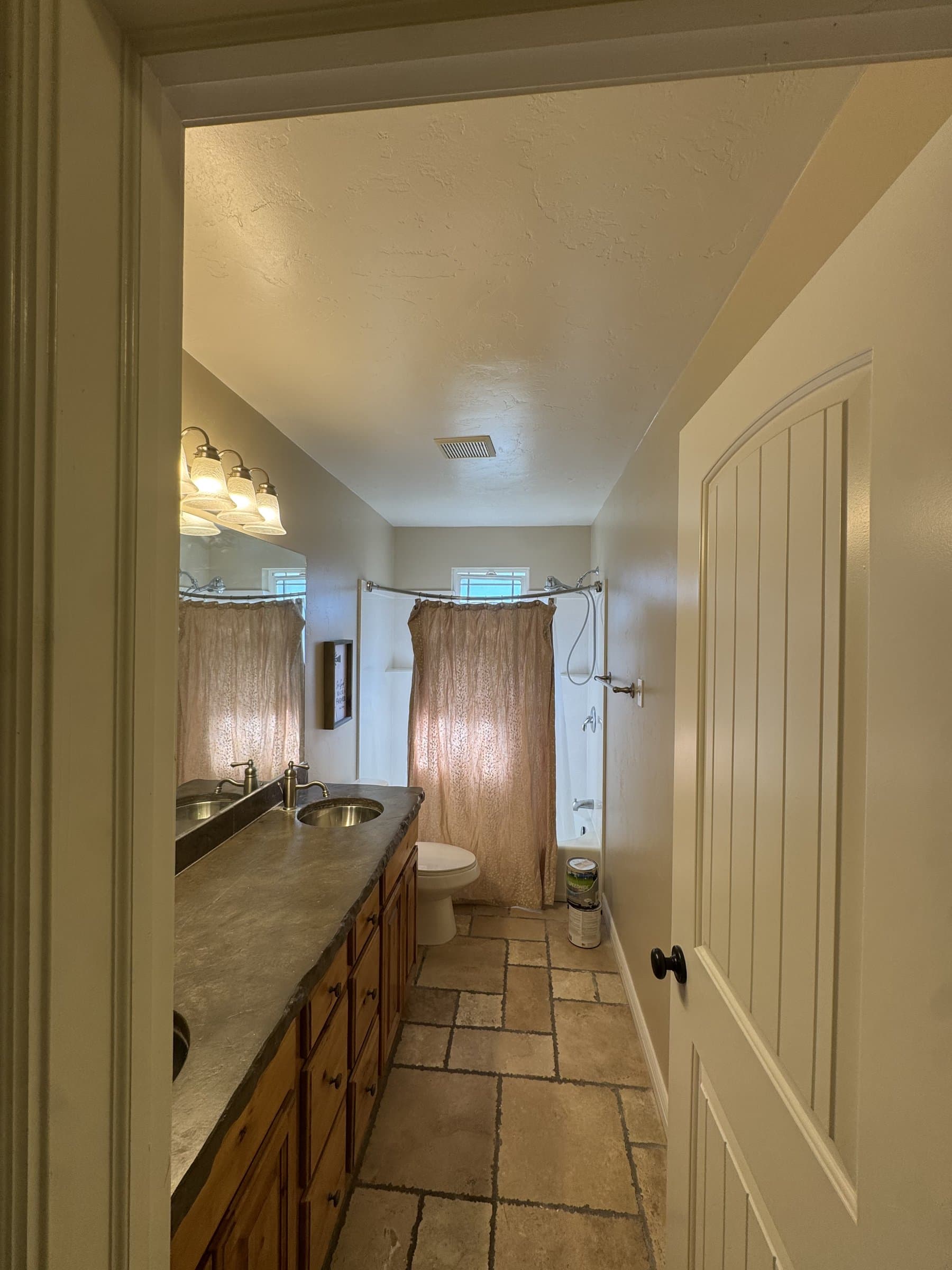 The same bathroom before work: an oak vanity with a poured concrete counter and two sinks, a tub and shower behind a curtain at the far end, travertine tile underfoot and cream walls throughout.