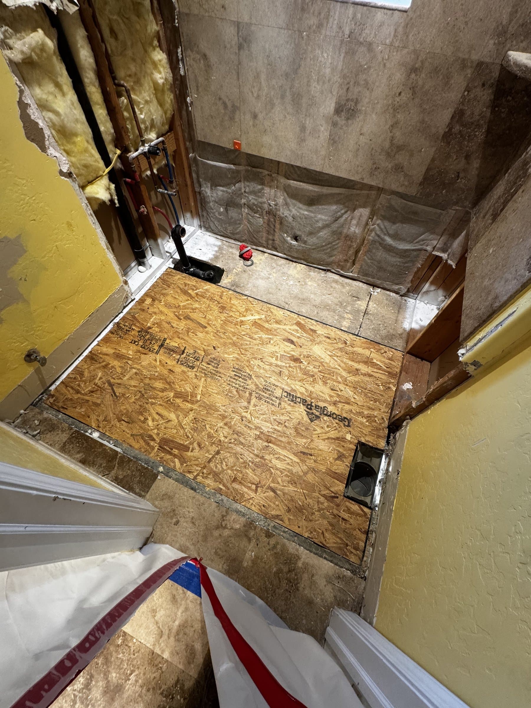 A shower stripped back to the structure: the pan lifted out down to a fresh plywood patch in the subfloor, supply pipes and the drain standing open, and the surrounding tile and drywall cut away to sound material.