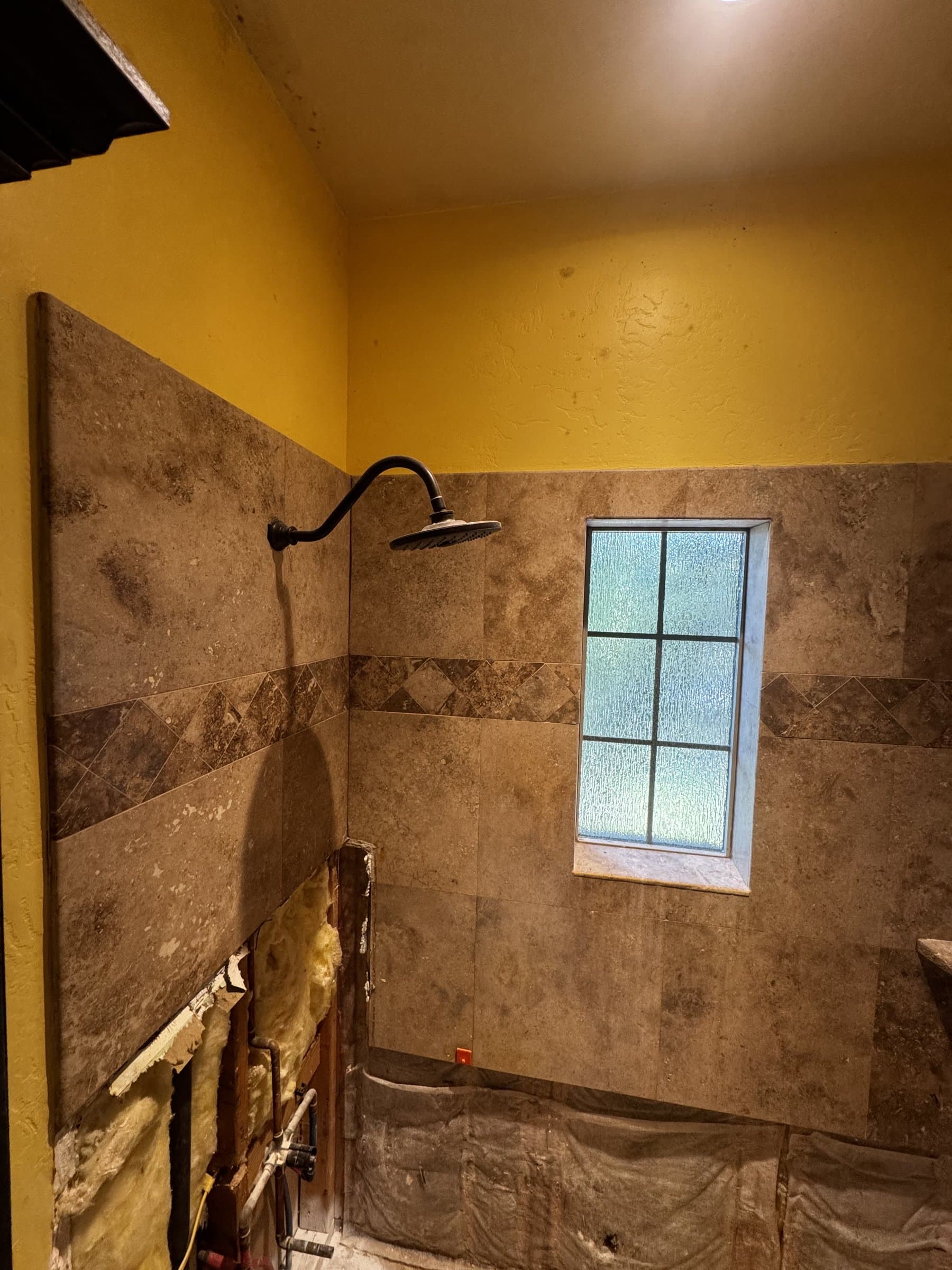 A travertine shower part-way through, with the tiled walls and their diamond border left sound and a section of the wet wall opened up beside the shower head to reach the valve behind it.
