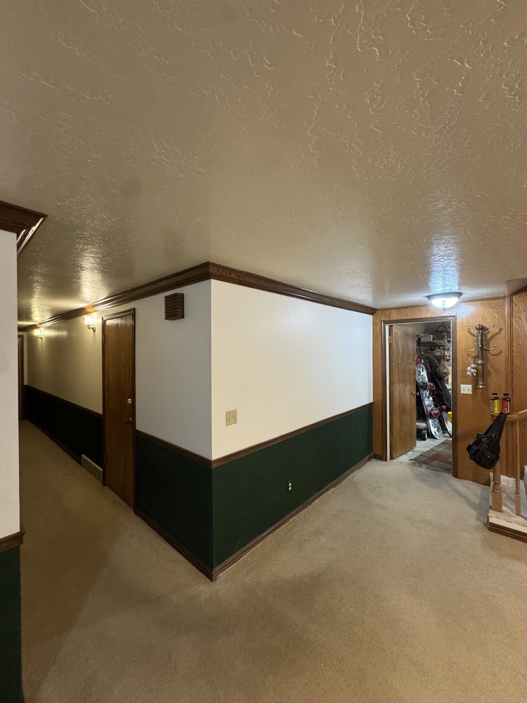 The basement hallway before work: a green painted dado under cream walls, dark stained chair rail and baseboard, oak doors and tan carpet running the length of the corridor.