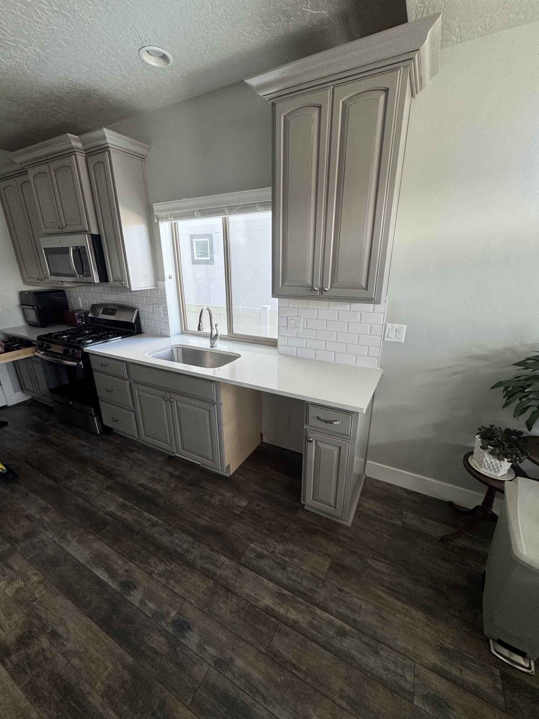 The finished kitchen looking down the run past the range and microwave, with gray cabinets, white quartz countertops, subway tile and dark plank flooring laid through to the far wall.