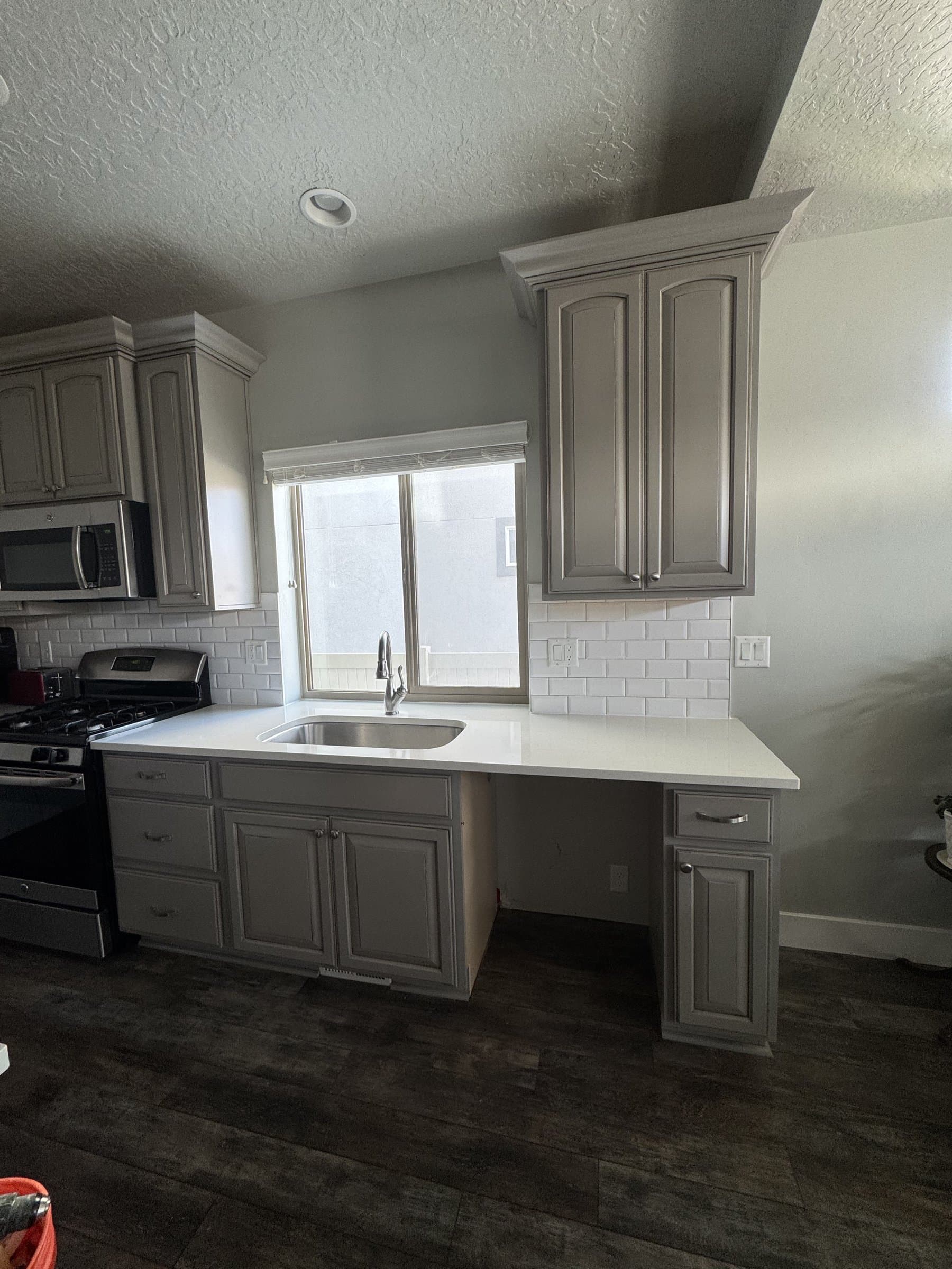 The finished kitchen sink run: gray paneled cabinets, a white quartz countertop, a stainless undermount sink under the window and white subway tile carried up to the underside of the wall units.
