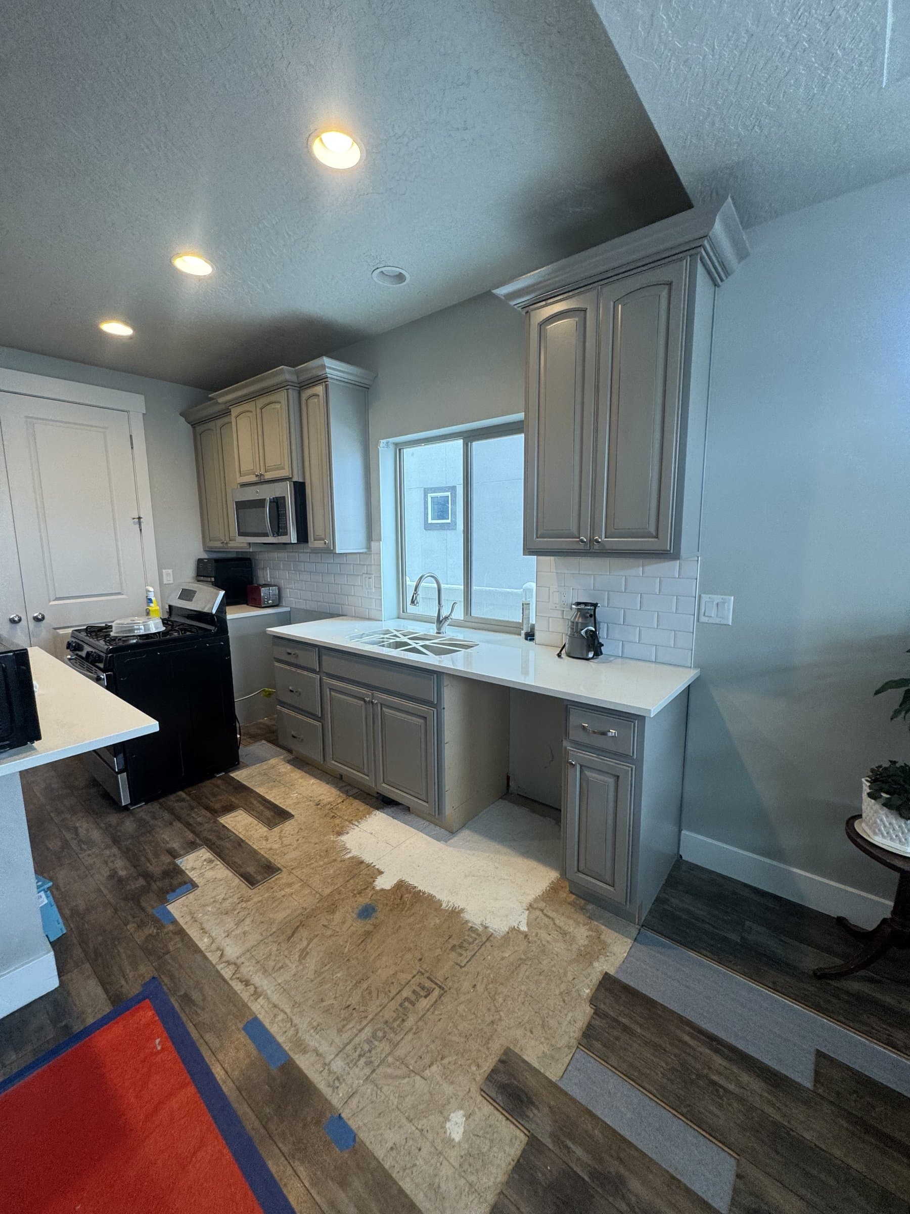 The kitchen with its gray cabinets, white quartz countertops and white subway tile backsplash all in place, photographed while the new plank flooring was still going down over the bare subfloor.