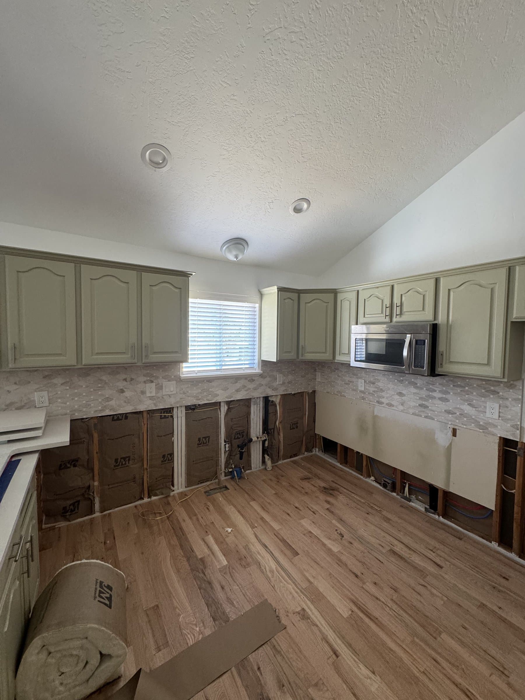 A kitchen part-way through: the sage green wall units and tiled backsplash still in place while the base cabinets are out, exposing insulated stud walls and open plumbing above newly laid oak flooring.
