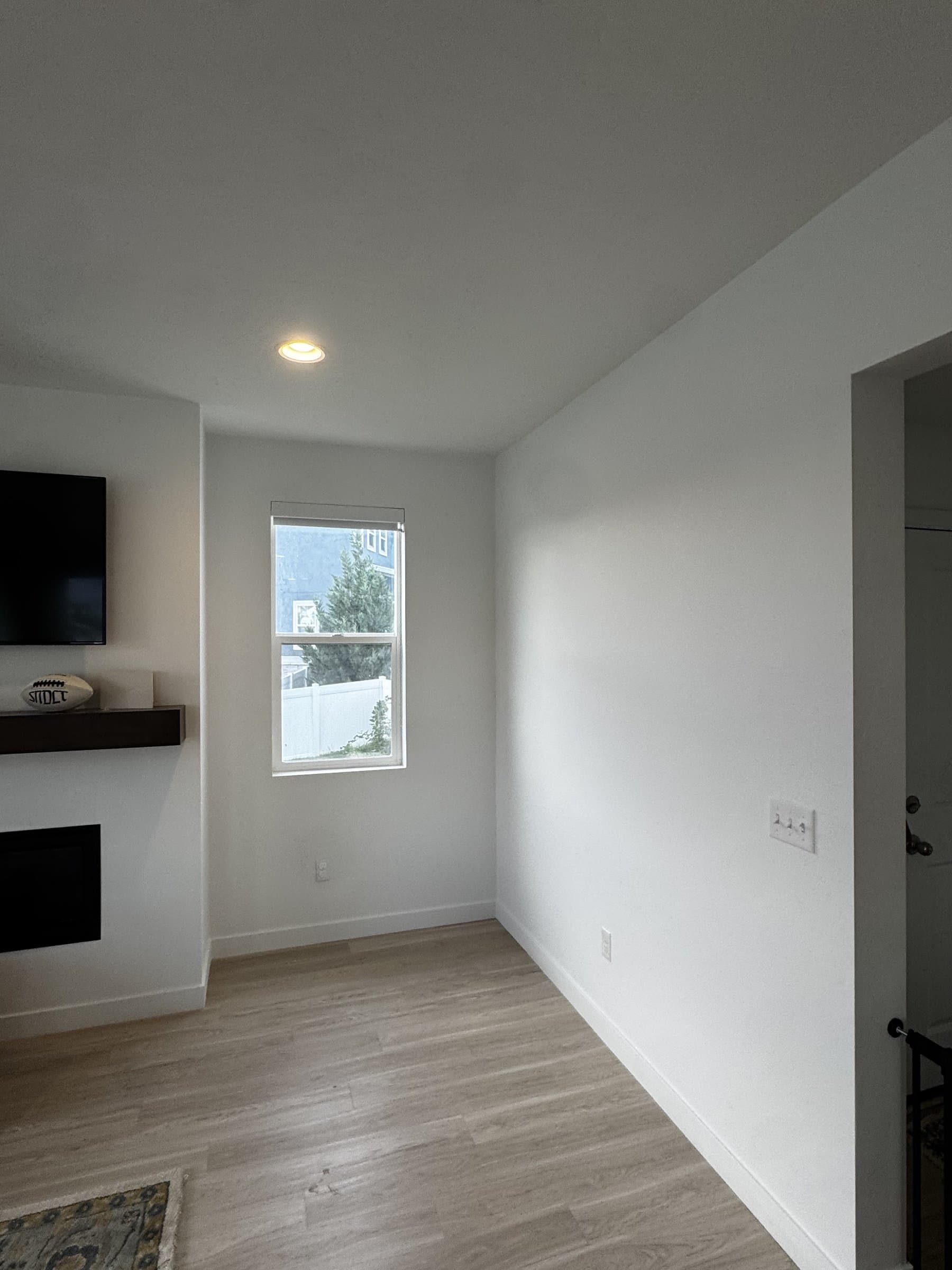 A finished living room corner painted flat white, with a modern fireplace under a dark floating mantel, pale plank flooring and a single window looking onto the fence line.
