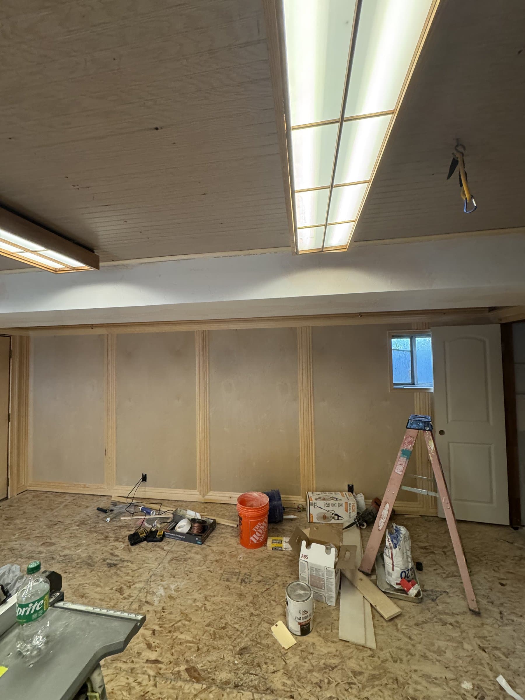 A 1970s basement room mid-project: the original tongue-and-groove wood ceiling and boxed fluorescent lights still overhead, new panel molding run across the walls unpainted, and buckets, a step ladder and offcuts across the bare subfloor.