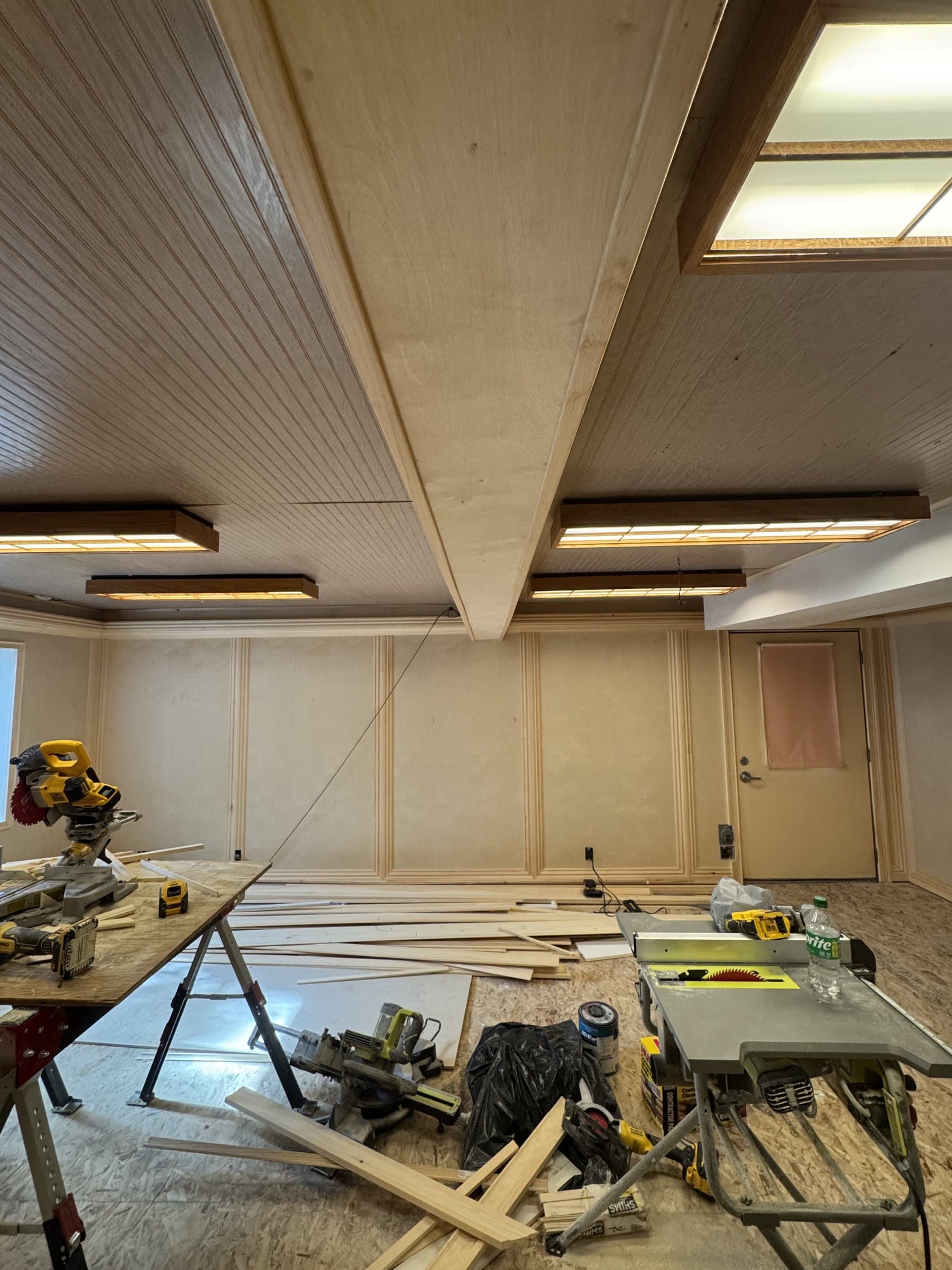 Trim day in the 1970s basement: a miter saw and a table saw set up on the bare subfloor, lengths of molding stacked and stickered ready to cut, and panel detail already run across the far wall under the original wood ceiling.